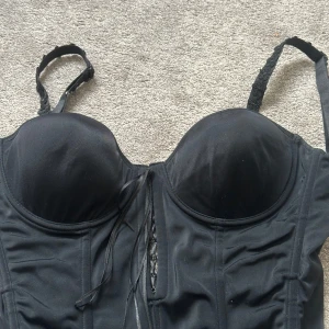 Svart korsett med snörning och spets - Snygg svart korsett med vadderade kupor och justerbara axelband dekorerade med blommig spets. Framtill finns snörning och hakar för att stänga korsetten. Materialet är mjukt och stretchigt, perfekt för att skapa en cool siluett. Passar dig som vill ha en edgy och feminin look.