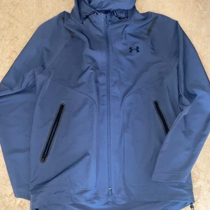 Vinna even svart färg Blå vindjacka från Under Armour  - Snygg blå vindjacka från Under Armour med huva och svarta detaljer. Jackan har dragkedja framtill, två fickor med svarta dragkedjor och justerbar huva med snörning. Perfekt för dig som gillar sportig stil och vill ha något funktionellt och stilrent.