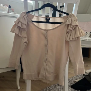 Rosa kofta med volang från H&M - Säljer en beige/rosa kofta från H&M med snygga volangdetaljer på axlarna och knappar framtill. Koftan har trekvartsärm och är tillverkad i mjuk bomull. 