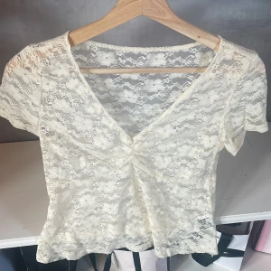 Spetsblus med v-ringning i ljus beige - Säljer en superfin ljus beige spetsblus med korta ärmar och v-ringning. Blusen har ett blommigt spetsmönster och är lätt transparent. Den har en snygg rynkning framtill som ger extra form. Perfekt för dig som gillar romantisk och feminin stil.