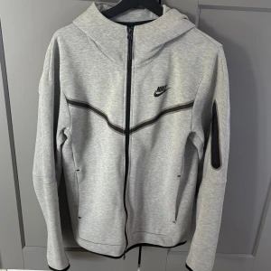Grå Nike Tech hoodie - Säljer min gråa nike tech hoodie som är i bra skick och utan några större defekter förutom det lilla man ser på bilden som knappt syns i verkligheten. 