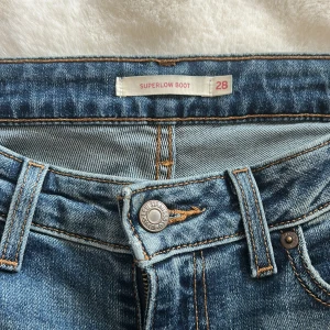Levi's Superlow Boot jeans blå - Snygga Levi's Superlow Boot jeans i klassisk blå tvätt med låg midja och bootcut-ben. Jeansen har fem fickor, orangea kontrastsömmar och Levi's ikoniska läderpatch bak. Perfekt för dig som gillar en avslappnad och cool stil.