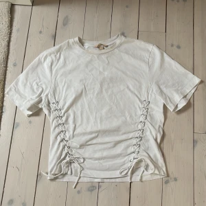 Vit t-shirt med snörning i sidan - Snygg vit t-shirt med cool snörningsdetalj på båda sidorna som ger en unik vibe. Modellen är kortärmad och har en klassisk rund hals. Perfekt att matcha med jeans eller shorts för en chill look. Materialet känns mjukt och skönt mot huden.