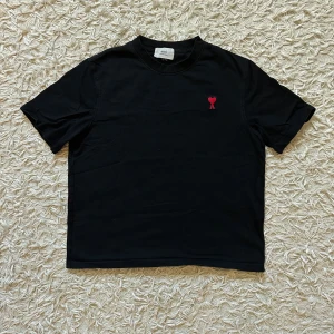 AMI PARIS- svart tshirt - Svart t-shirt från AMI med klassisk passform och korta ärmar. På bröstet finns ett broderat rött hjärta med bokstaven A under, och baktill vid nacken är AMI broderat ton-i-ton. Tillverkad i mjuk bomull, perfekt för en clean och stilren look.
