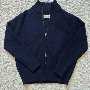 MM tjock marinblå ikoniska tröja - Säljer en marinblå stickad kofta från MM (ma1son margie1a, kan ej skriva märket i beskrivningen då annonsen tas bort av nån anledning- men ni förstår nog) med ribbad struktur och hel dragkedja framtill. Tröjan har hög krage och långa ärmar, samt de ikoniska vita stygn på ryggen. Tillverkad i Italien och passar perfekt för dig som gillar stilrena designer. Den är helt ny aldrig använd med tagsen som medföljer, obs den är väldigt tjock- heavyweight och håller en varm. 