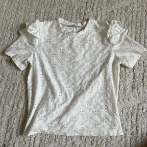 Vit blus med volang från Zara - Supersöt vit blus från Zara med korta ärmar och volangdetaljer på axlarna. Blusen har broderat hålmönster över hela och rund halsringning. Aldrig använd 