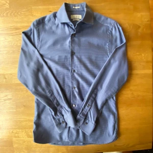 Blå skjorta från The Tailoring Club - Snygg blå skjorta från The Tailoring Club i premium bomull. Skjortan har ett diskret rutigt mönster, klassisk krage och knappar framtill. Långärmad modell med stilrena manschetter. Perfekt för dig som gillar en clean och modern look.