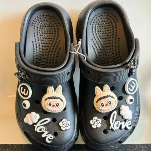Platå tofflor med dekorationer  - Helt nya och oanvända svarta platå slip in skor med gulliga Labubu dekorationer  Dom är tillverkade i gummi, så dom tål vatten Det är en lätt & bekväm modell Sulans höjd8cm Ytterlängd ca 27cm Innerlängd ca 22,8-23cm Storlek 36-37