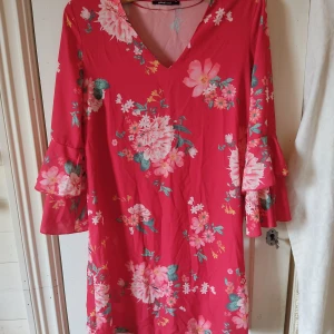 Blommig sommarklänning - En röd klänning från Gina Tricot med blommigt mönster i rosa och grönt. V-ringad hals och trekvarts ärmar med volanger längst ner. Materialet känns lätt och passformen är vid och luftig så den passar bra som en sommarklänning.