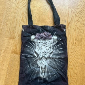 Svart shoppingväska Van Asch med tryck - Cool svart shoppingväska från Van Asch med ett detaljerat tryck av en dekorerad tjurskalle och lila rosor. Väskan har långa handtag och är gjord i bomull.