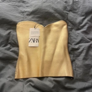 Guldig tubtopp från Zara - Supersnygg guldig tub-topp från Zara i storlek S. Toppen är axelbandslös med en stilren bandeau-form och har en diskret dragkedja i sidan. Materialet är glansigt och ger en lyxig känsla, perfekt för att sticka ut. Grym att matcha med jeans eller kjol för en trendig look.