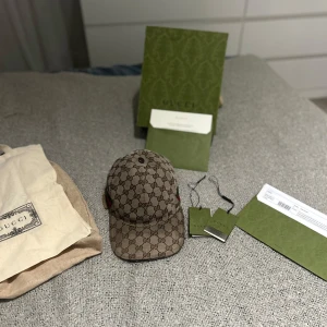 Gucci keps med GG-mönster, storlek S - Säljer en snygg beige keps från Gucci med det klassiska GG-monogrammet över hela kepsen. Kepsen har en böjd skärm och gröna/röda ränder på sidan. Tillverkad i bomull och polyester, perfekt för dig som vill ha en lyxig streetstyle-accessoar.  fås med kvitto box dustbag, med andra ord alla tillbehör. Säljer den billigt pga ganska sliten. Pris ej hugget i sten. MVH/ damskiii