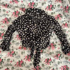Blommig croppad blus från H&M Divided - Supersöt svart croppad knytblus från H&M i strl 32 med daisys på. Blusen har långa ärmar, krage, knappar och knytning framtill!💞