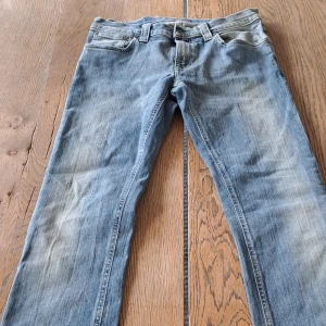 Blå jeans från Nudie Jeans, 28/32 - Säljer ett par klassiska blå jeans från Nudie Jeans i storlek 28/32. De har en snygg tvättad look med ljusa partier, raka ben och fem fickor. Jeansens material är bomull och de har Nudie Jeans logga på bakfickan och läderpatch i midjan. Jeansen är i väldigt bra skick, de är lite skrynkliga pga de har legat i garderoben ett tag utan användning.