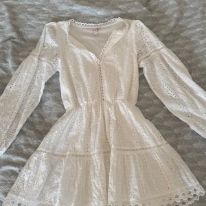 Vit spetsklänning från Nelly - Superfin vit kortklänning från Nelly med broderade spetsdetaljer och v-ringad hals. Klänningen har långärmade puffärmar och markerad midja som ger en snygg siluett. Perfekt för sommarens alla tillfällen, med romantiska volanger och genombrutet mönster.