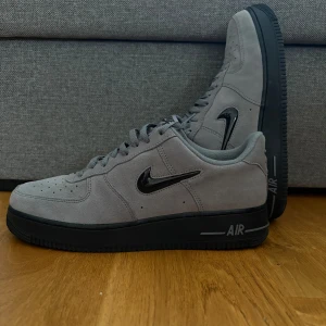 Nike Air Force 1 grå mocka sneakers - Nike Air Force 1 sneakers i grå mocka med svart sula och blank svart swoosh på sidan. Klassisk låg modell med snörning och rund tå. Snygg kontrast mellan den mjuka ovandelen och den robusta svarta sulan. Perfekt för dig som gillar stilrena och moderna sneakers.