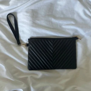 Svart quiltad kuvertväska med lösbar handtag - Snygg svart kuvertväska med quiltad yta i chevronmönster på framsidan och slät baksida. Väskan har en avtagbar handledsrem i svart och guldfärgade metalldetaljer. Stängs med dragkedja upptill och har ett rymligt fack för dina essentials. Perfekt att bära i handen             Måtten på väskan  Längd 27cm Bred 1 cm Rem längd 22cm Höjd 17,5cm