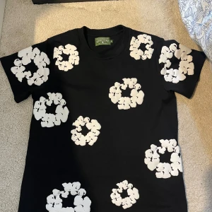 Svart t-shirt med vita blommor Denim Tears - Svart t-shirt från Denim Tears med stora vita blommiga tryck över hela plagget, både fram och bak. Rund hals och korta ärmar. Materialet är mjuk bomull och passformen är normal. Perfekt statement-plagg för dig som gillar unik design.