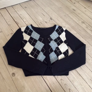 Argyle kofta från Brandy Melville - Snygg kofta från Brandy Melville med argylemönster i svart, ljusblått och vitt. V-ringad med knappar framtill och ribbade muddar. Mjuk stickad kvalitet som är perfekt för lager-på-lager. Långärmad och croppad passform som ger en trendig look. Aldrig blivit använd helt ny men utan prislappen kvar 