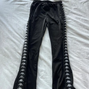 Svarta Kappa träningsbyxor med knappar - Svarta träningsbyxor från Kappa med klassiska vita loggor längs båda sidorna. Byxorna har elastisk midja och tryckknappar längs benen för att enkelt kunna öppnas. Snygg sportig look och skön passform, perfekta för chill eller träning.