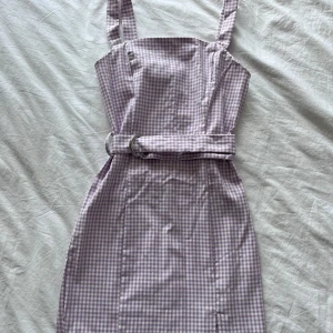 Lila rutig kortklänning med bälte - Supersöt lila och vit rutig kortklänning med axelband och tillhörande bälte i midjan. Klänningen har en slim passform och en liten slits framtill. Perfekt för dig som gillar en feminin och trendig look med retrovibbar.