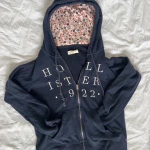 Mörkblå hoodie från Hollister med blommig huva - Snygg mörkblå hoodie från Hollister med vit text på framsidan och dragkedja. Insidan av huvan har ett blommigt mönster i färger som orange, rosa och turkos. Tröjan har långa ärmar, snörning vid huvan och mjukt material som känns skönt mot huden.