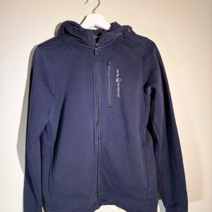 Sail Racing Hoodie - Snygg mörkblå hoodiejacka från Sail Racing med dragkedja framtill och en bröstficka med logga. Jackan har huva med snörning och långa ärmar, perfekt för chill dagar. Materialet är mjukt och känns skönt mot huden.
