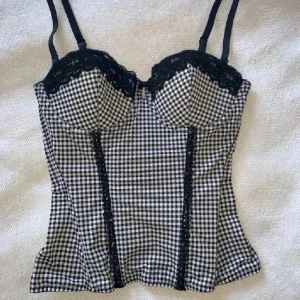 Svartvit rutig korsett med spets - Supersnygg korsett-topp i svartvitt rutigt mönster med tunna axelband och svarta spetsdetaljer längs kuporna och framtill. Toppen har en figurnära passform och en liten rosett mellan kuporna. Perfekt för dig som vill sticka ut med en cool och trendig look.