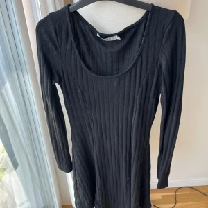 Svart ribbad klänning från NA-KD - Snygg svart ribbad klänning från NA-KD med lång ärm och rundad halsringning. Modellen är kort och har en fin passform som framhäver figuren. Perfekt för dig som gillar stilrena och enkla plagg med lite edge.