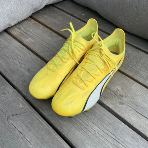 Puma Ultra gula fotbollsskor - Säljer ett par Puma Ultra fotbollsskor i en riktigt fräsch gul färg med svarta detaljer och vit sidopanel. Skorna har snörning och är tillverkade i lätt syntetmaterial för maxad speed på planen. Sulan har fasta dobbar för gräs och Puma-logga både bak och under.
