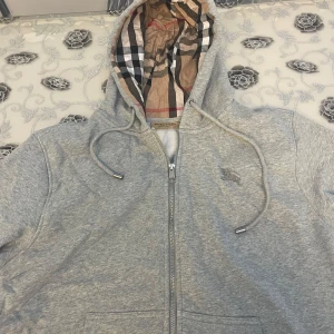 Grå Burberry kofta/tröja - Säljer en grå hoodie från Burberry med klassiskt rutigt foder i huvan och broderad logga på bröstet. Tröjan har snörning i huvan, praktisk dragkedja och är tillverkad i mjuk bomull. Perfekt för dig som gillar stilrena och lyxiga detaljer. Passar bättre som L