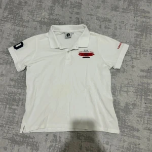 Vit Hang Ten t-shirt med tryck - Vit t-shirt från Hang Ten med klassisk krage och korta ärmar. Framsidan har ett rött surfbräde-tryck med texten 'Hang Ten', och ena ärmen har en stor mörkblå '60'. På andra ärmen står 'HANG TEN' i rött. Skön bomullskvalitet och sportig vibe.