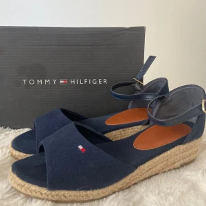 Marinblå sandaler från Tommy Hilfiger - Snygga marinblå sandaler från Tommy Hilfiger med öppen tå, justerbar ankelrem och flätad platåsula i rep. Ovandelen är i textil med diskret logga framtill. Perfekta för sommaren och ger en fräsch look till alla outfits.