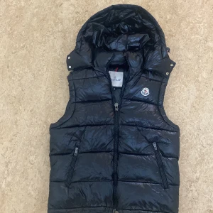 Svart dunväst från Moncler med huva - Svart, glansig dunväst från Moncler med avtagbar huva och två dragkedjefickor framtill. Västen har quiltad design och klassisk Moncler-logga på bröstet. Perfekt för lager-på-lager och håller dig varm med äkta dunfyllning.