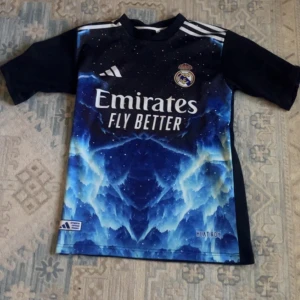  Helt ny Real Madrid Adidas fotbollströja svart o blå  - Snygg Real Madrid fotbollströja från Adidas med coolt blått och svart mönster framtill, klubbmärke och sponsortryck. Kortärmad modell i lätt funktionsmaterial, perfekt för träning eller match. Ikoniska vita Adidas-ränder på axlarna.