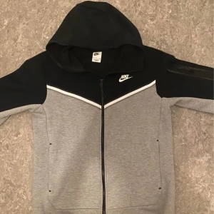 Nike Tech Fleece hoodie grå/svart - Nike Tech Fleece hoodie i grått och svart med vit detalj över bröstet. Jackan har huva, hel dragkedja och fickor på sidorna samt en extra ficka med dragkedja på ärmen. Perfekt för chill eller streetwear.