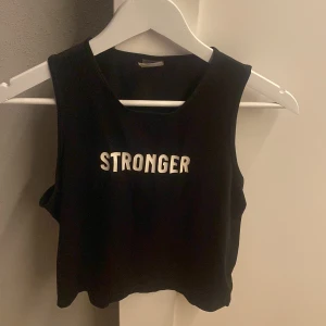 Svart croppad linnetopp från Stronger - Svart croppad linnetopp från Stronger med vit text framtill. Toppen är ärmlös och har en loose passform, perfekt för träning. Tillverkad av 90% återvunnen polyester och 10% elastan för en skön och stretchig känsla.