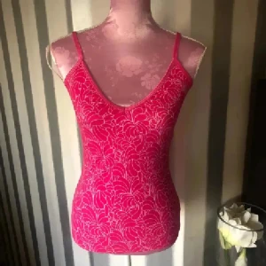 Rosa blommig linnetopp med smala axelband - Snygg rosa linnetopp med blommigt mönster och smala axelband. Toppen har en v-ringad hals och är figurnära i modellen. Perfekt för dig som gillar färg och vill sticka ut lite extra. Materialet känns lätt och bekvämt mot huden. Inbyggd bh. Storlek m men lite mindre i storleken 