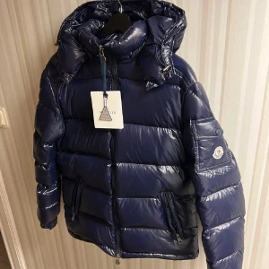 Mörkblå pufferjacka från Moncler - Snygg mörkblå pufferjacka från Moncler med glansig finish och bruna detaljer på insidan. Jackan har huva, dragkedja framtill och två sidofickor med dragkedja. Klassisk Moncler-logga på ärmen och coolt serietryck på insidan. Perfekt för kalla dagar.SKRIV FÖR FLER BILDER FÖR DE BLEV LITE SUDDIGA! Loggan är lite suddig på bilderna men skriv så skickar jag fler bilder 