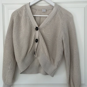 Beige stickad kofta från Lindex - Snygg beige stickad kofta från Lindex med grovstickad struktur och tre svarta knappar framtill. Koftan har långärmade ärmar och breda ribbade muddar vid nederkant och ärmslut. Inte använd många gånger!💕