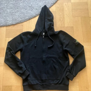 Svart hoodie från Stone Island - Svart hoodie från Stone Island med klassisk logotyp-patch på vänster ärm. Tröjan har huva med snörning, hel dragkedja framtill och två stora fickor. Materialet är mjuk bomull och passformen är normal. Perfekt för dig som gillar streetwear och snygga detaljer. Storlek L men passar mer som M