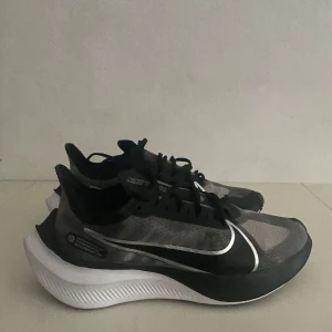 Nike Zoom Gravity svarta sneakers - Nike Zoom Gravity sneakers i svart och grått med vit sula och stor vit swoosh på sidan. Lätt och ventilerande mesh-överdel, snörning och platt sula med coolt mönster undertill. Perfekta för dig som gillar sportig och modern stil. Dom har använts 2 gånger utomhus men är i helhet mycket rena och stadiga