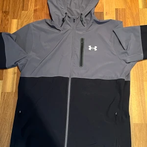 Under Armour vindjacka grå/svart - Snygg tvåfärgad vindjacka från Under Armour i grått och svart. Jackan har huva med snörning, hel dragkedja och praktiska fickor med dragkedja. Materialet är lätt och syntetiskt, perfekt för utomhusaktiviteter. Stilren logga på bröstet och modern passform.