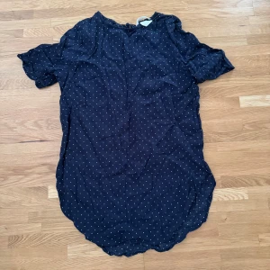 Mörkblå prickig kortklänning strl 34 - Söt mörkblå kortklänning med vita prickar och rundad nederkant. Klänningen har korta ärmar med lätt volang och knäppning bak i nacken. Perfekt för dig som gillar en lekfull och chill stil.