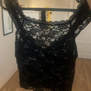 Svart spetsig linnetopp med rosett - Supersnygg svart linnetopp i spets med genombrutet mönster och en liten rosett framtill. Toppen har breda axelband och vacker vågig kant upptill. Perfekt för dig som gillar detaljer och vill ha en feminin vibe på outfiten.