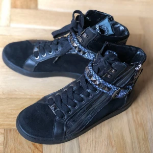 Svarta high-top sneakers Michael Kors - Snygga svarta high-top sneakers från Michael Kors med coola detaljer som snörning, dragkedja och ett mönstrat band med pyramidnitar runt ankeln. Skorna har en mix av mocka och skinn, samt ett reptilmönster som ger extra edge. Skick: Nästan nya, använd några gånger. 