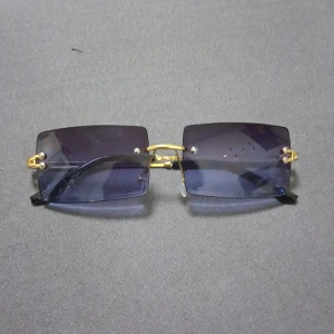 Rektangulära blå solglasögon med gulddetaljer - Snygga rektangulära solglasögon med blåtonade linser och guldfärgade metallbågar. Skalmarna är svarta och har en tunn, elegant design. Glasögonen har näskuddar för extra komfort och en modern, minimalistisk look som sticker ut.