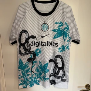 Inter Milan fotbollströja Nike - Inter Milan Dri-Fit fotbollströja från Nike i vitt med svarta och turkosa detaljer. Tröjan har korta ärmar, svart krage och grafiskt mönster med ormar och växter. Materialet är lätt och andas, perfekt för träning eller match.