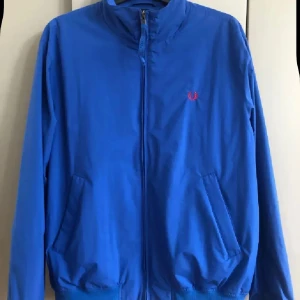 Blå jacka från Fred Perry M  - Snygg blå jacka från Fred Perry med dragkedja och två fickor framtill. Jackan har ett litet rött Fred Perry-logo broderat på bröstet och ribbade muddar vid midja och ärmslut. Perfekt för dig som gillar stilren och sportig look.