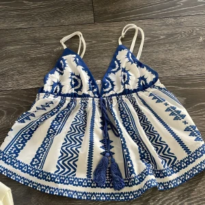 Vit och blå mönstrad linnetopp - Supersöt linnetopp med tunna axelband och v-ringning. Toppen har ett blått och vitt etnoinspirerat mönster och knytband med tofsar i midjan. Perfekt för varma dagar och ger en chill vibe till din outfit.